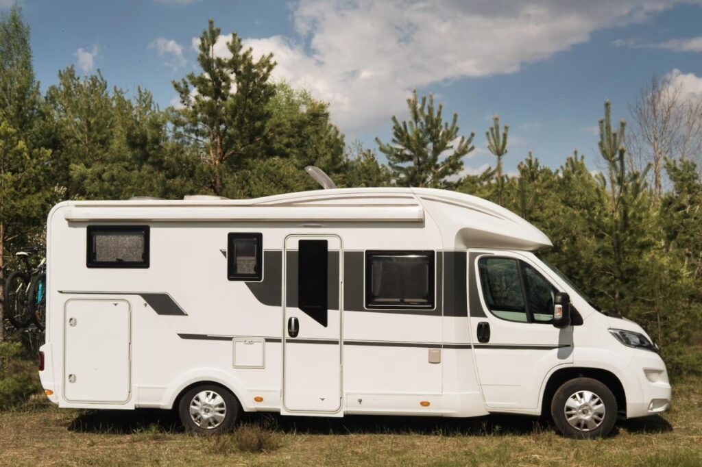 A white camper van parked in a forest clearing, surrounded by pine trees, showcasing its side profile and exterior design. Ideal for travel enthusiasts.